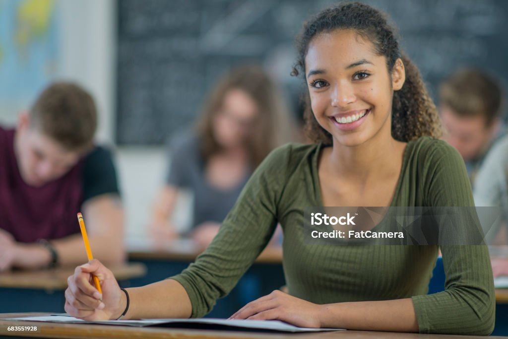 A multi-ethnic group of university students are indoors at school. They are in a classroom. They are wearing casual clothing. They are writing a standardized test. A girl of African descent is in front. She is holding a pencil and smiling at the camera.